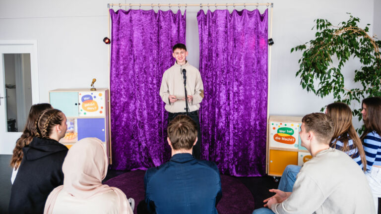Ein Jugendlicher steht auf einer Bühne vor einem lilafarbenen Vorhang und springt zu Jugendlichen, die vor ihm sitzen und von hinten zu sehen sind